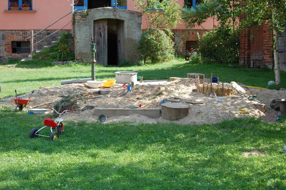 Sandfläche mit Spielzeug, Holzstämmen und einer grünen Handpumpe auf der Wiese